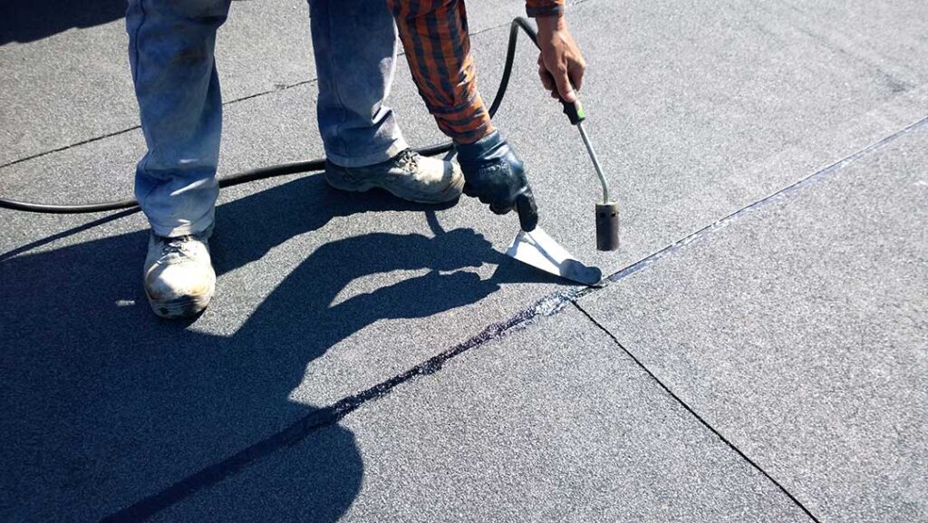 A homeowner wearing gloves uses a roof rake to clear leaves from a residential roof on a sunny day.