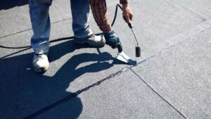A homeowner wearing gloves uses a roof rake to clear leaves from a residential roof on a sunny day.